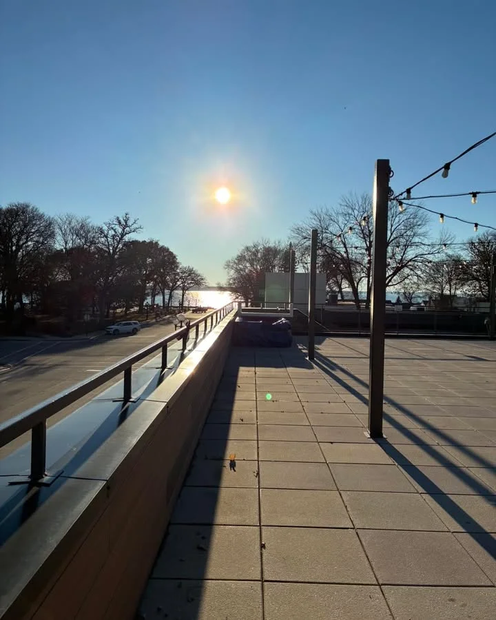 A clear day view from a rooftop, featuring a sun setting over the horizon with trees lining the background.