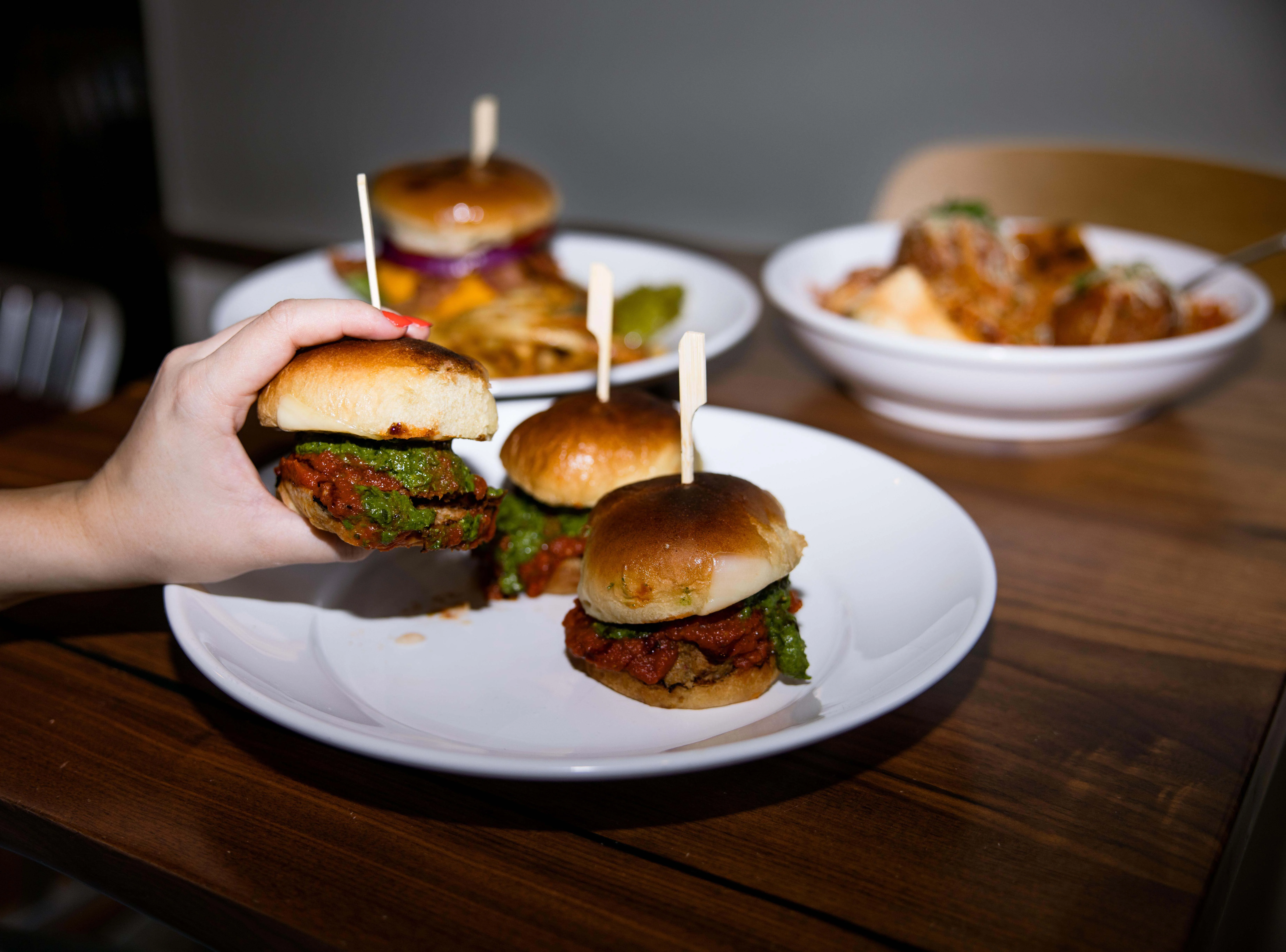 A hand holding a slider with two patties and green sauce, with other sliders and a pasta dish in the background.