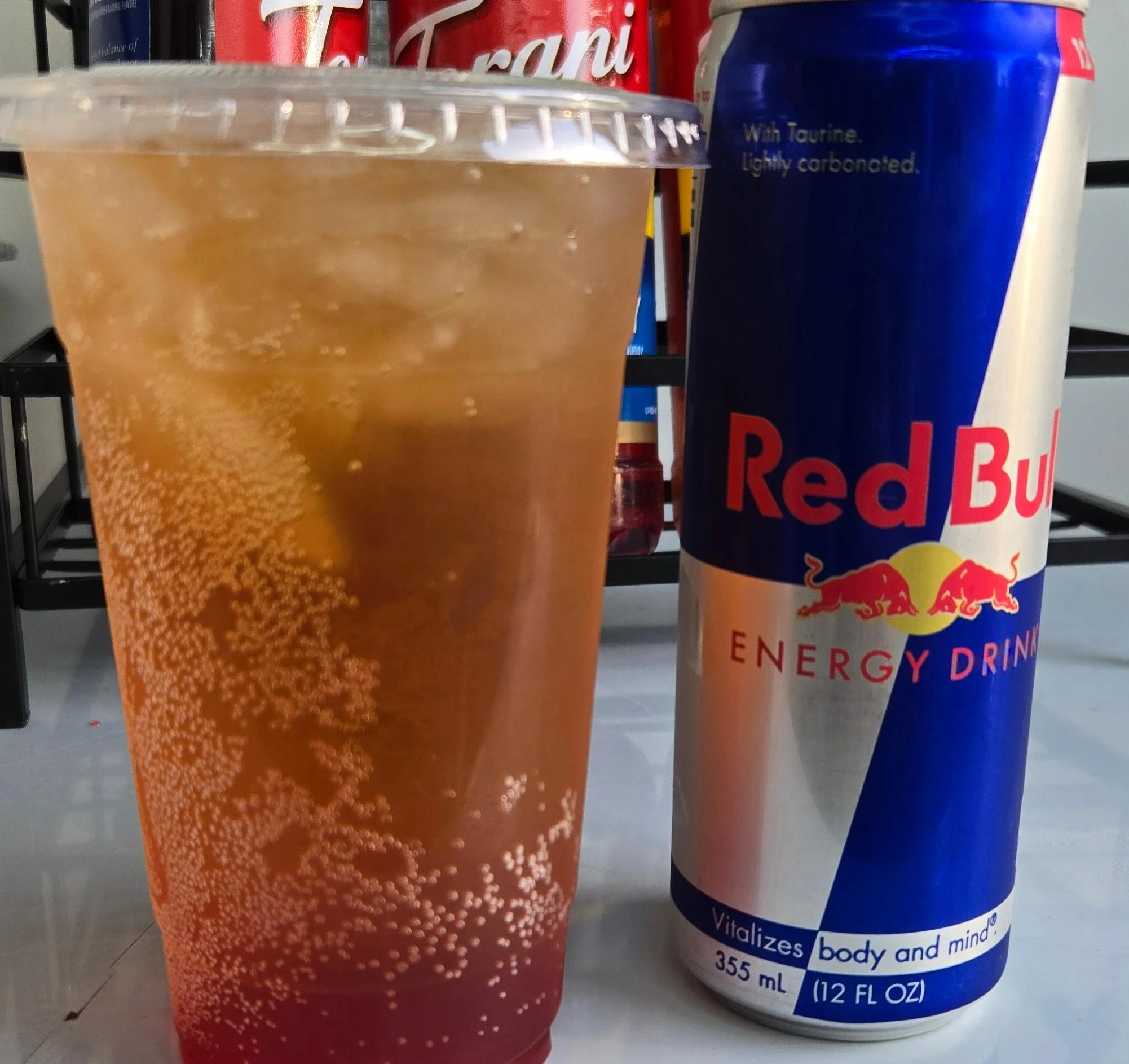 A clear cup filled with a fizzy brown beverage and ice, alongside a can of energy drink. The cup is decorated with bubbles, indicating carbonation.