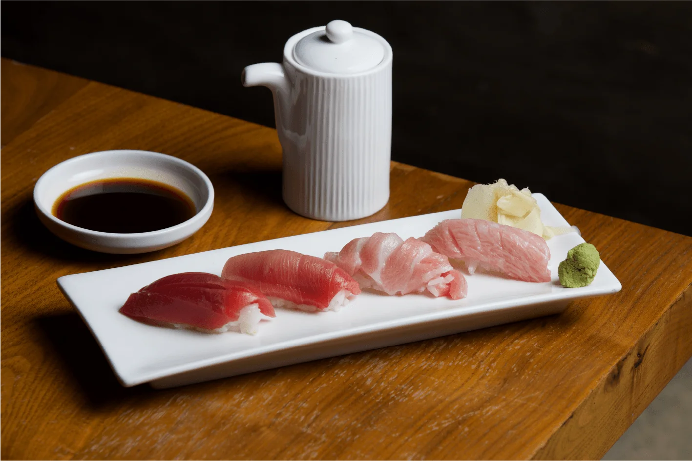 A selection of four distinct cuts of tuna nigiri sushi arranged on a rectangular white plate, accompanied by a small pile of pickled ginger, wasabi, a small dish of soy sauce, and a white ceramic sake or soy sauce pitcher on a wooden table.