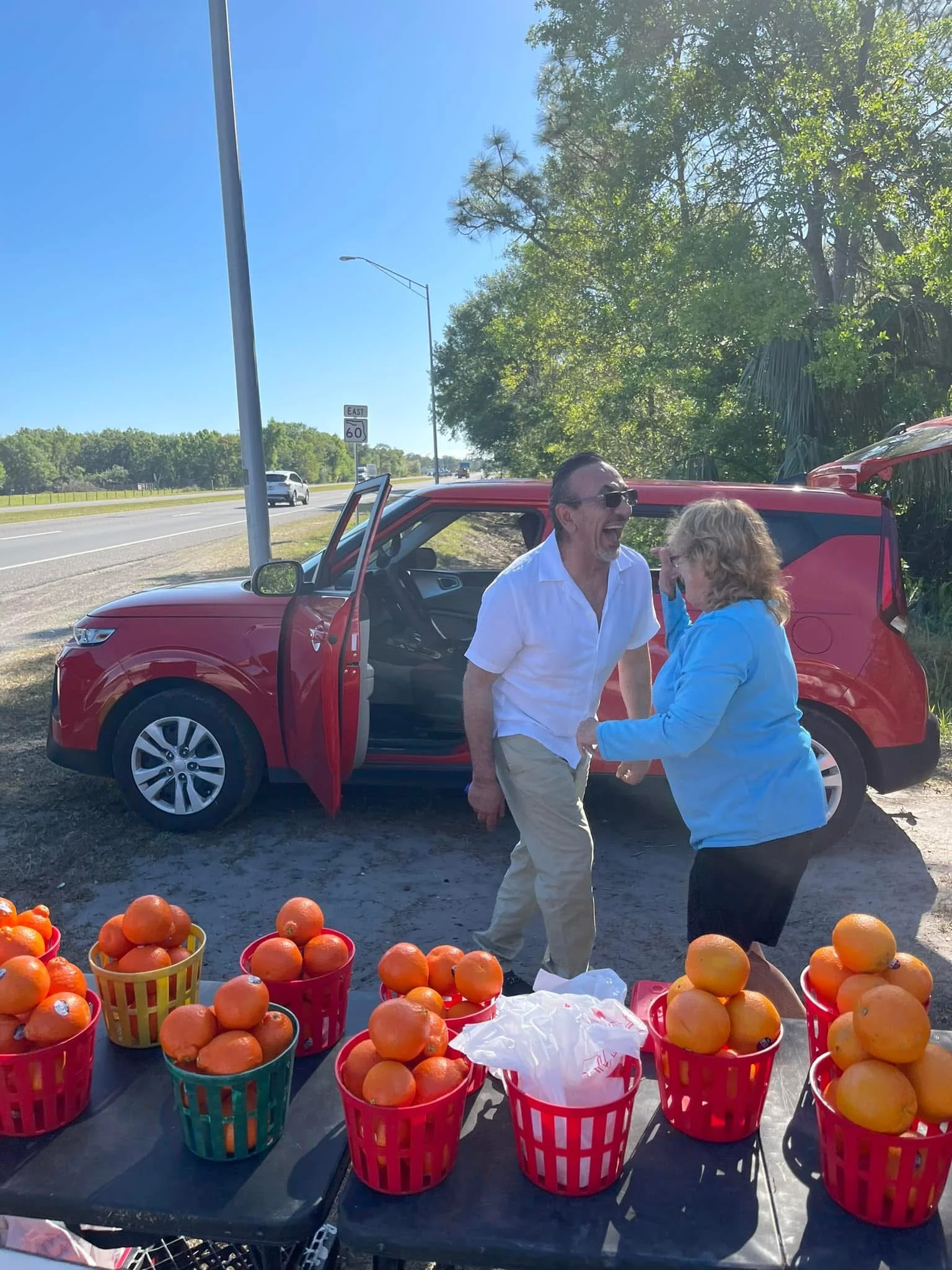 Two people laughing by a roadside fruit stand with oranges