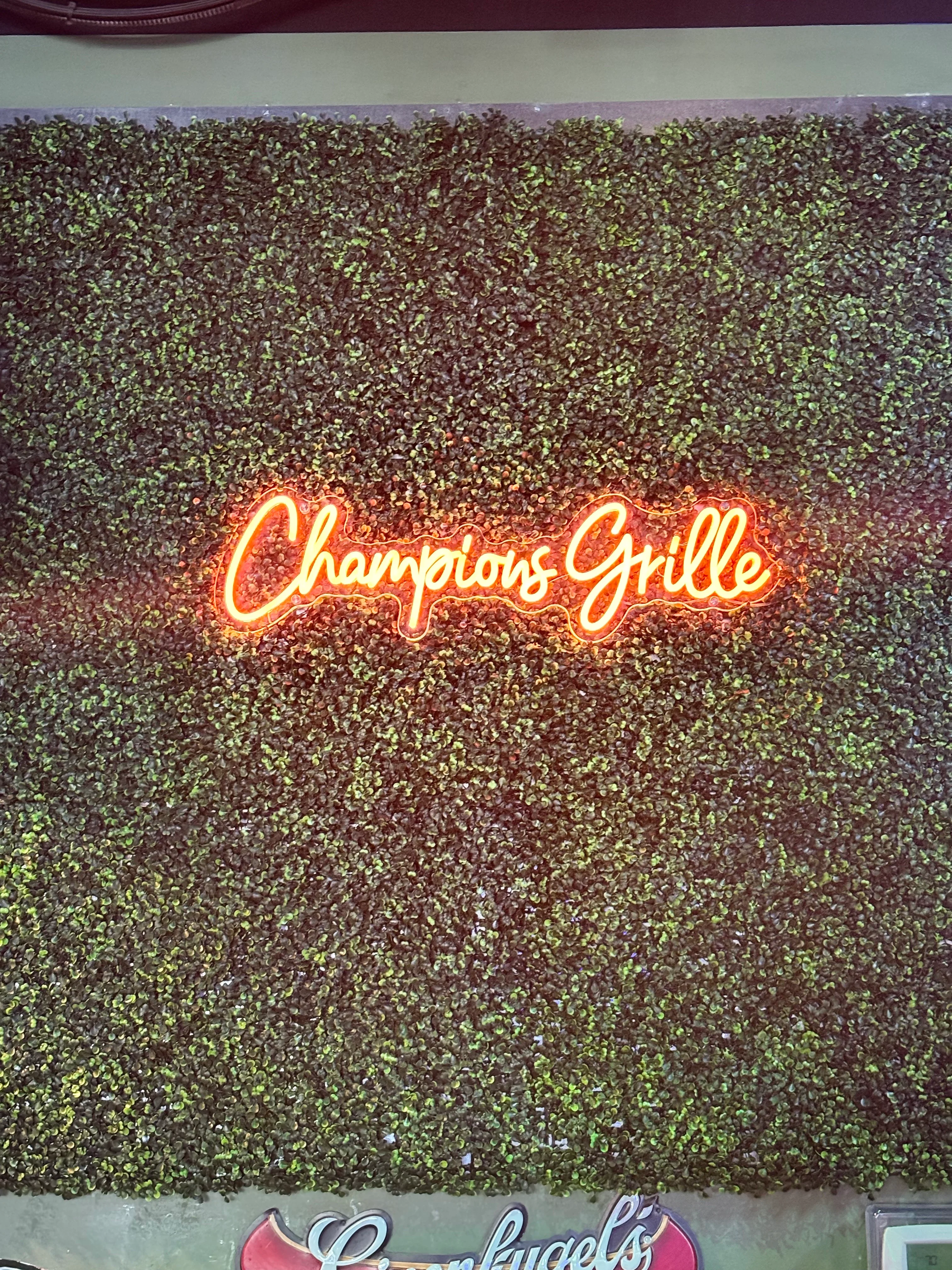 A neon sign displaying the words 'Champions Grille' against a backdrop of green foliage.
