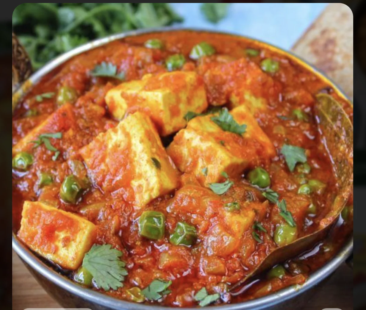 A bowl of rich tomato-based curry with paneer cubes and green peas, garnished with fresh herbs.