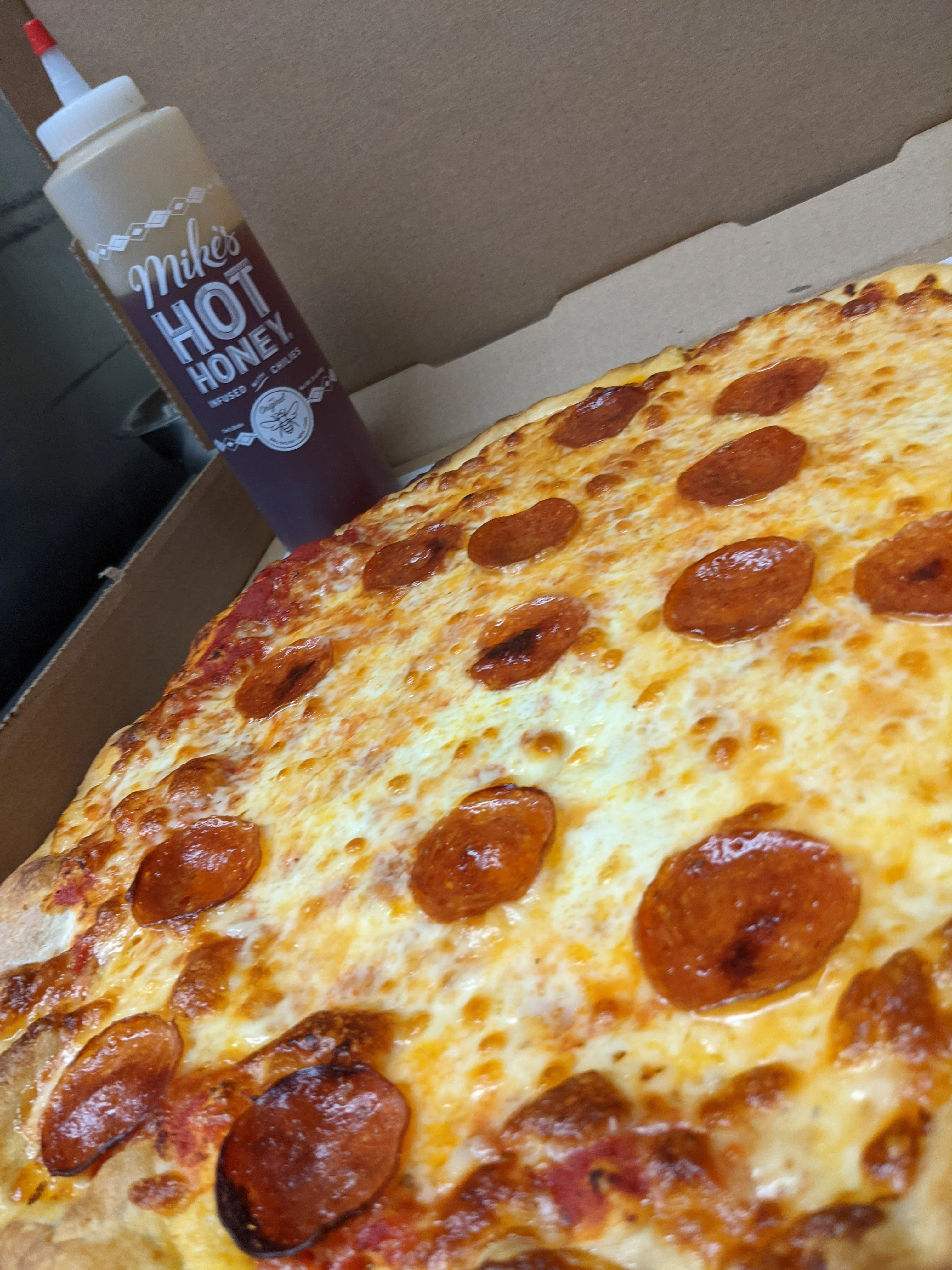A box containing a large pepperoni pizza next to a bottle of hot honey sauce.