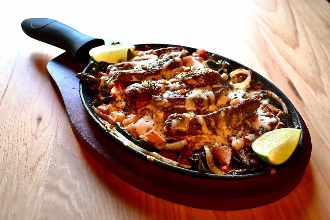 A sizzling plate featuring grilled meat with melted cheese, onions, and diced tomatoes, garnished with lime wedges.