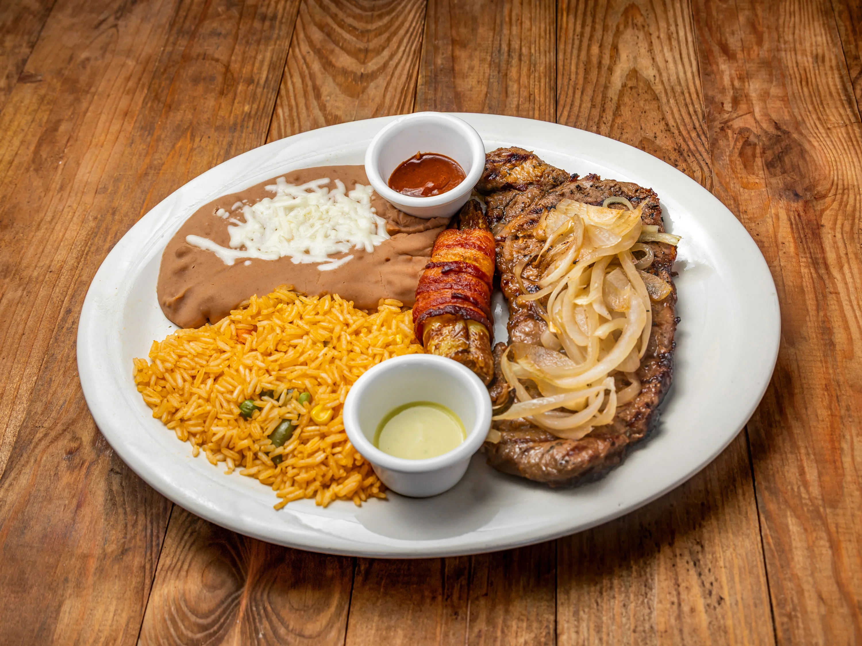 A plate featuring grilled steak topped with onions, accompanied by refried beans, yellow rice, and a wrapped sausage. The dish includes small bowls of sauce and lime.