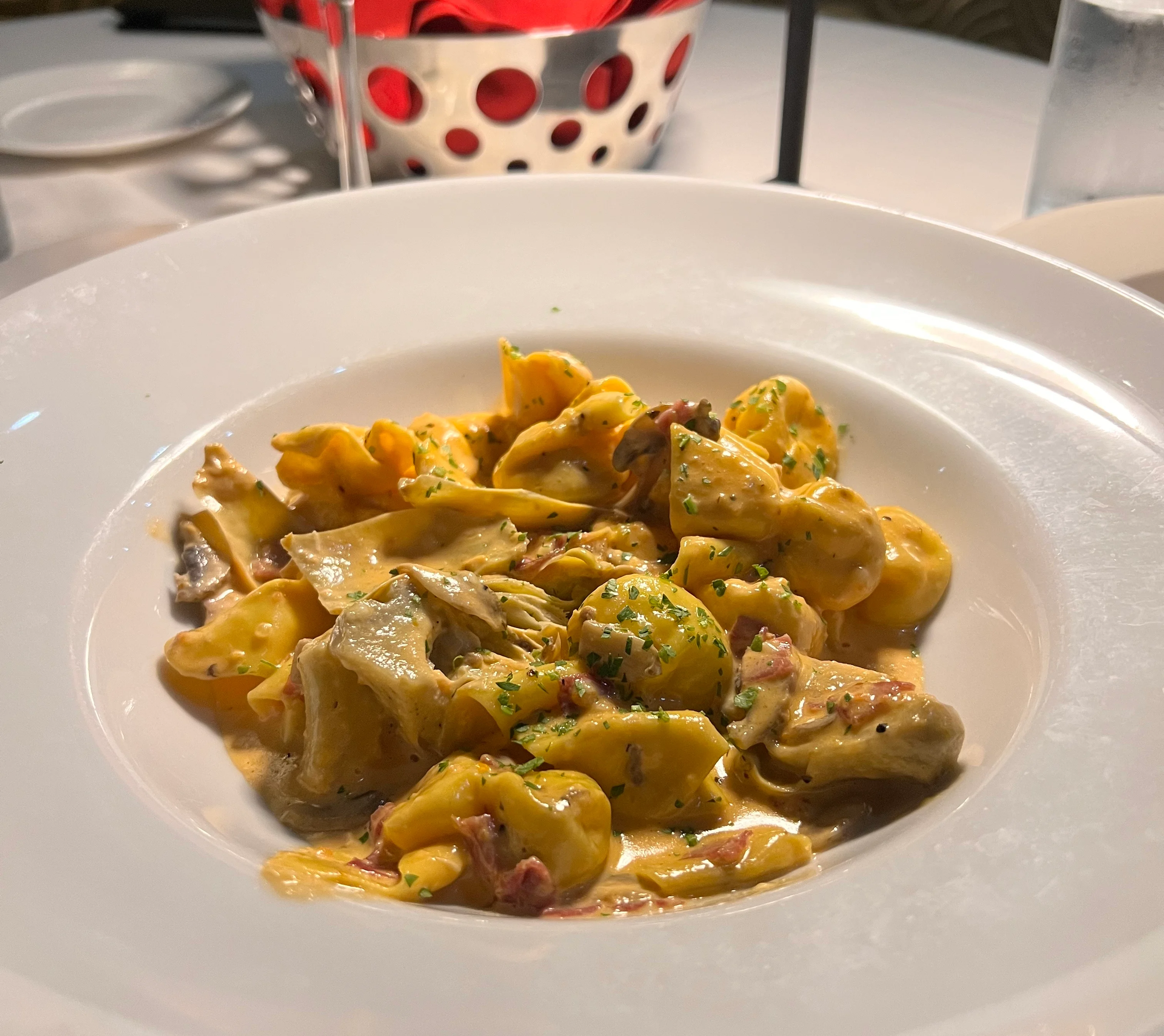 A close-up of a creamy tortellini pasta dish garnished with herbs, served in a white bowl on a restaurant table with a cocktail, bread basket, and water glass in the background.