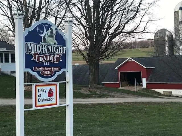 A sign for a family dairy farm, indicating its history since 1836, with a backdrop of farm buildings and trees.