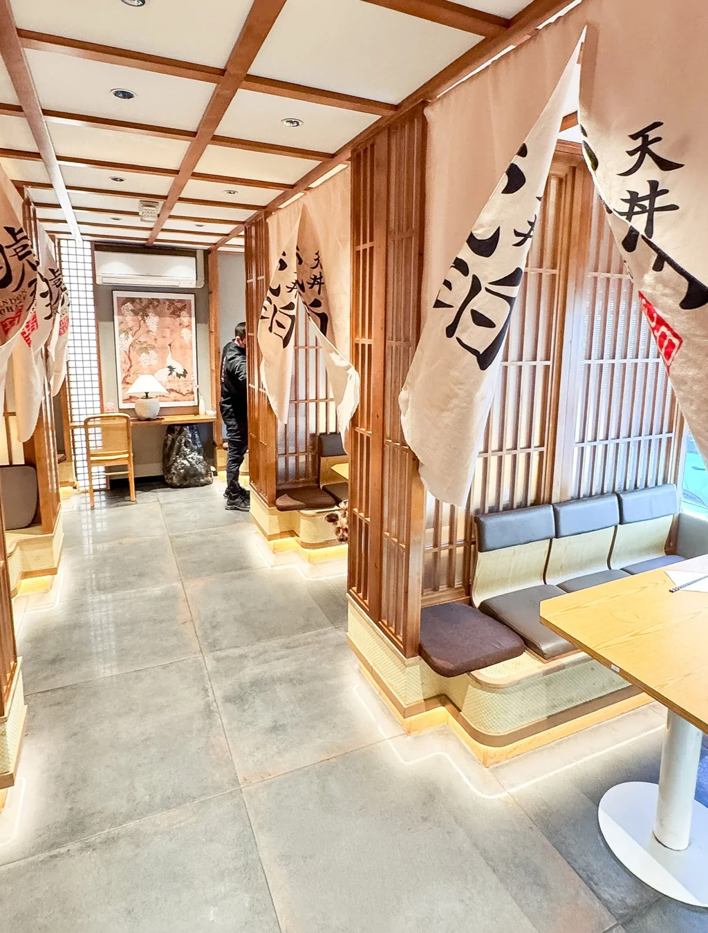 A bright, wide-angle interior view of a contemporary Japanese restaurant hallway. The space features light wood paneling, clean stone flooring, private booth seating shielded by hanging white fabric banners, and soft ambient lighting reflecting off the surfaces.