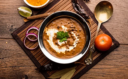 A bowl of lentil soup garnished with cream and cilantro. The dish is served on a wooden tray alongside spices, a lime, and a small orange.