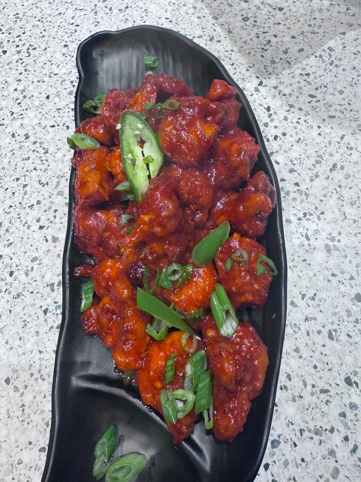 A black dish filled with spicy cauliflower florets coated in a vibrant red sauce, garnished with green onions and slices of green chili.