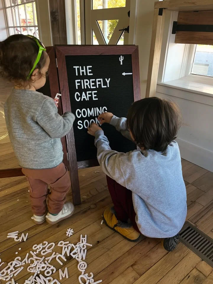 Two children are arranging letters on a sign board that reads 'THE FIREFLY CAFE COMING SOON.' The scene is set in a cozy space with wooden flooring.