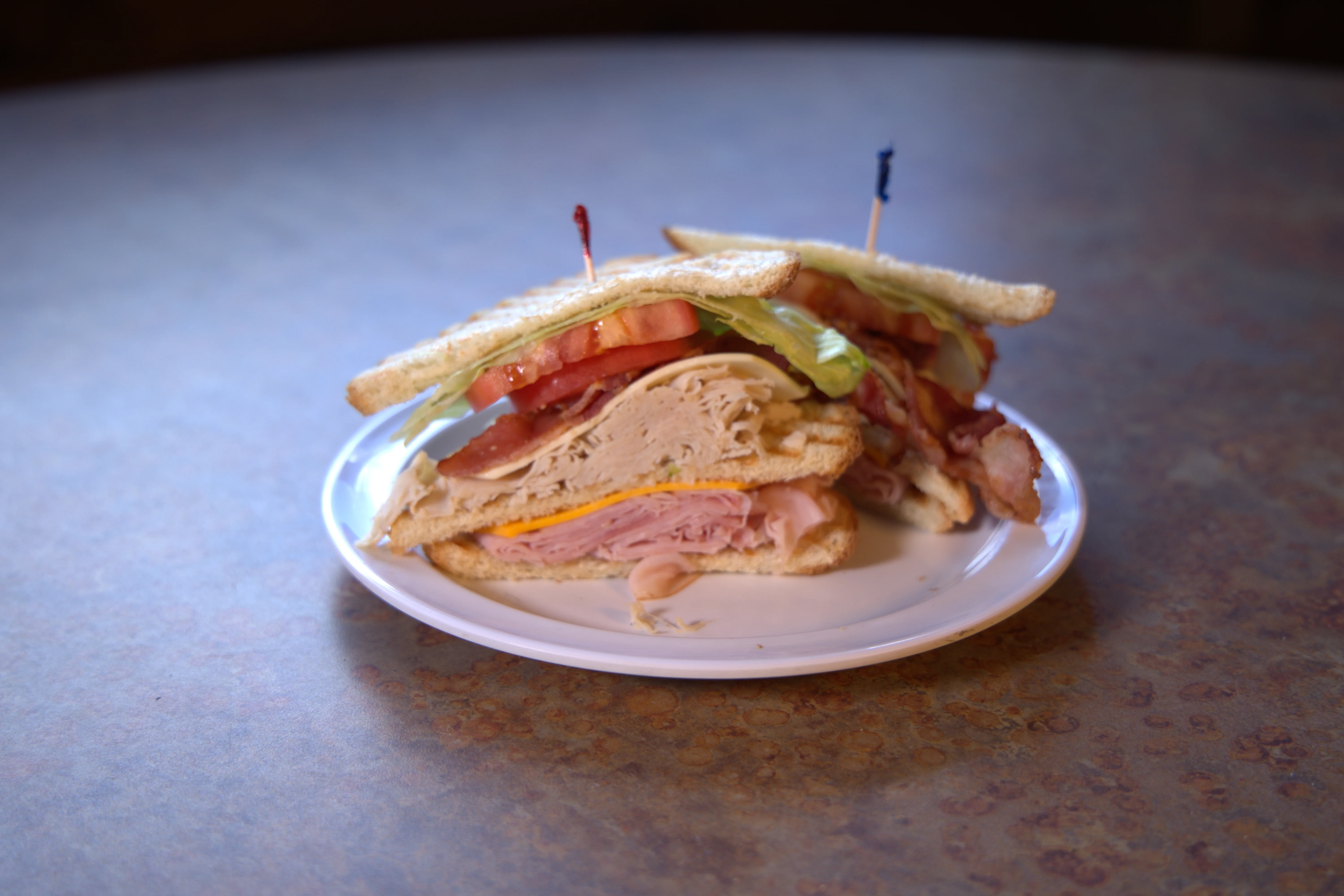 A stacked sandwich with layers of turkey, ham, bacon, lettuce, and tomatoes, garnished with toothpicks on a plate.