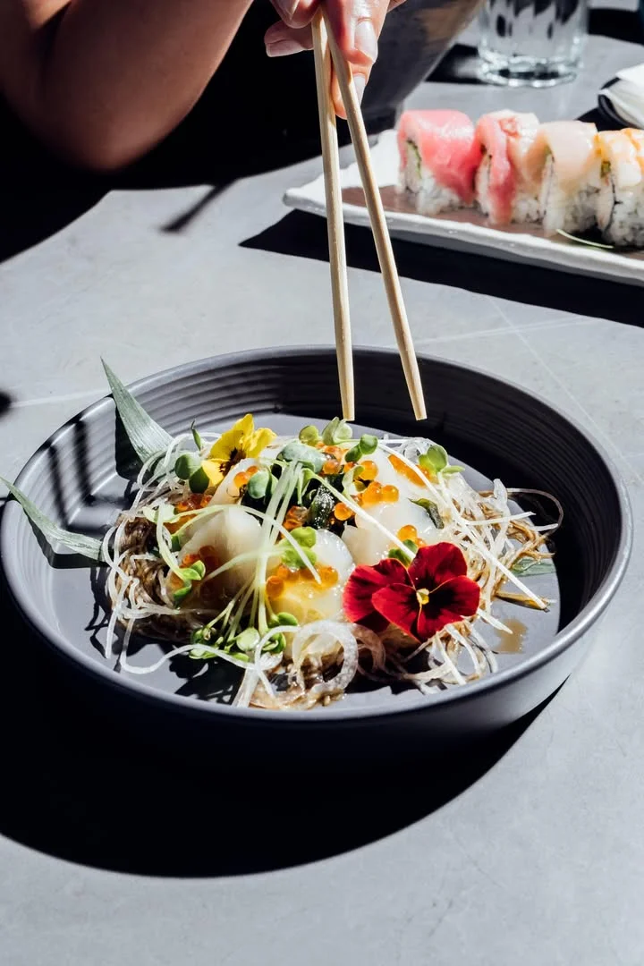 A decorative bowl filled with seafood, garnished with sprouts, flowers, and served with chopsticks, alongside sushi in the background.