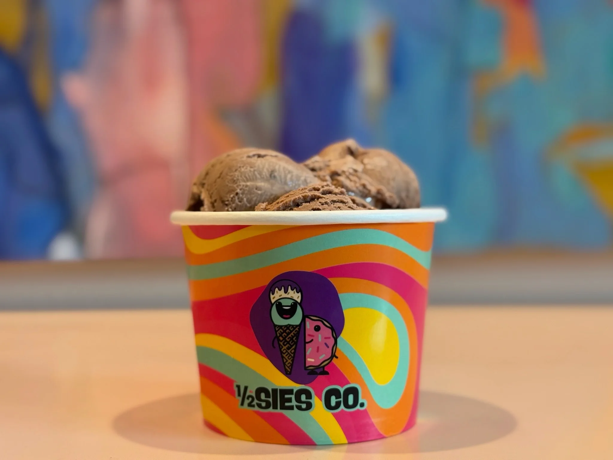 A colorful ice cream cup filled with scoops of chocolate ice cream. The cup features a vibrant design with playful colors and an illustration.