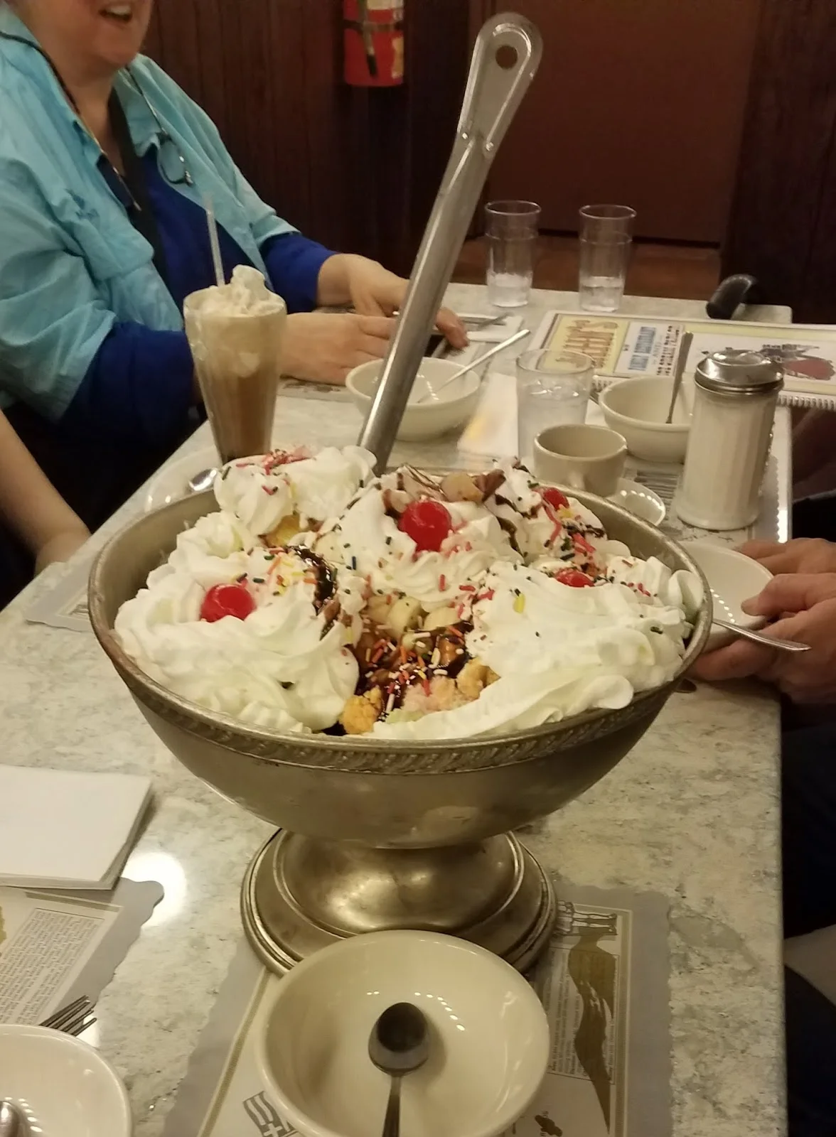 A large bowl of ice cream topped with whipped cream, cherries, sprinkles, and syrup, served at a diner.
