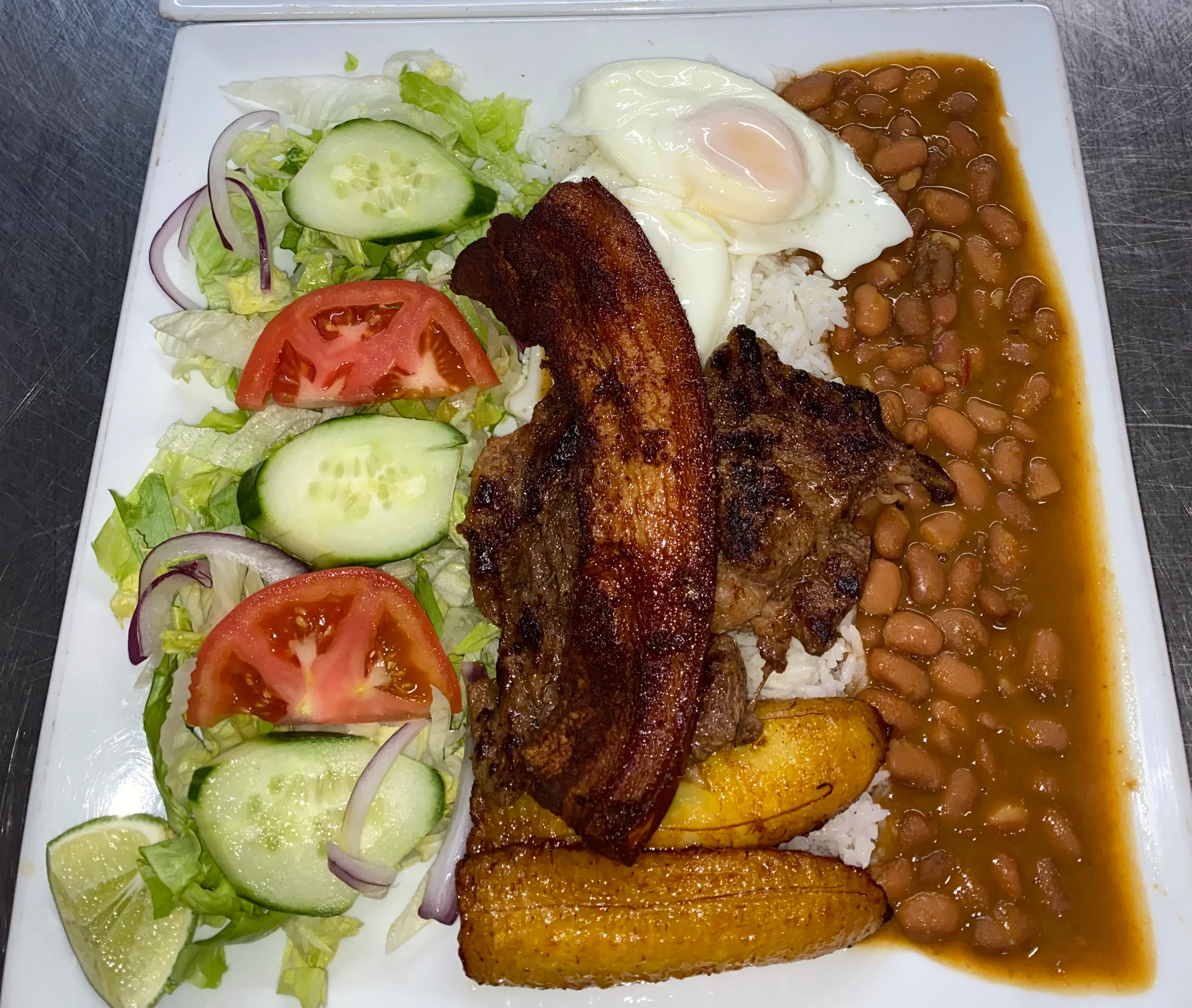 A hearty dish with a fried egg, grilled meats, rice, beans, plantains, and a fresh vegetable salad on a white plate.