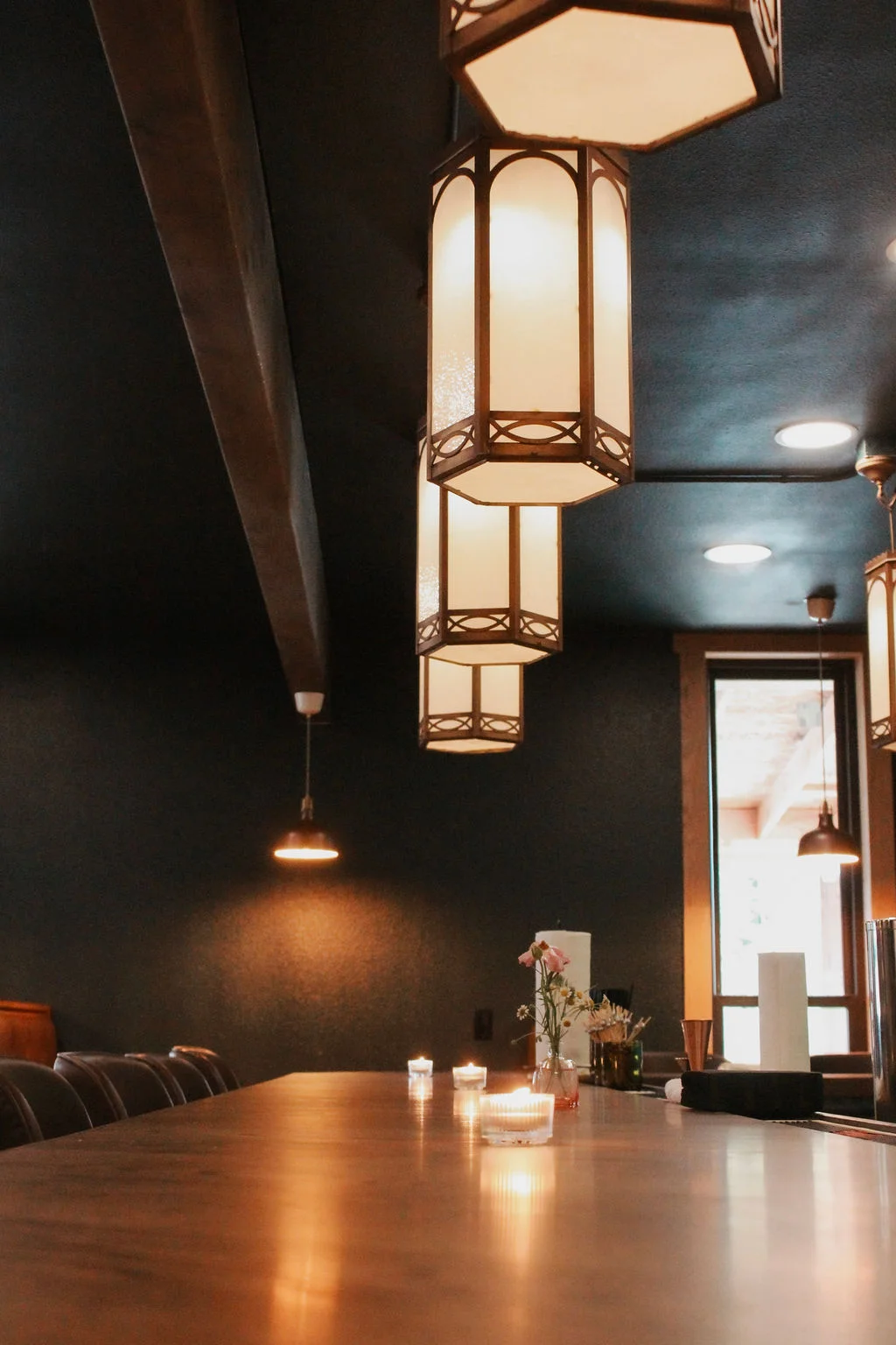 A modern dining area featuring a long wooden table with candles and floral arrangements. Soft lighting hangs from the ceiling, creating a warm atmosphere.