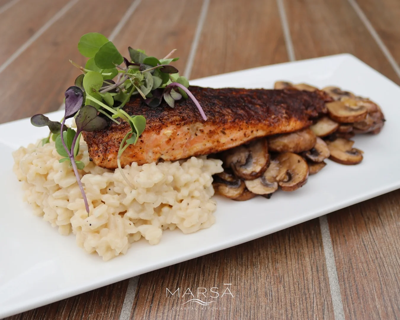 A plated dish consisting of salmon fillet seasoned and served on a bed of creamy risotto with sautéed mushrooms and garnished with microgreens.
