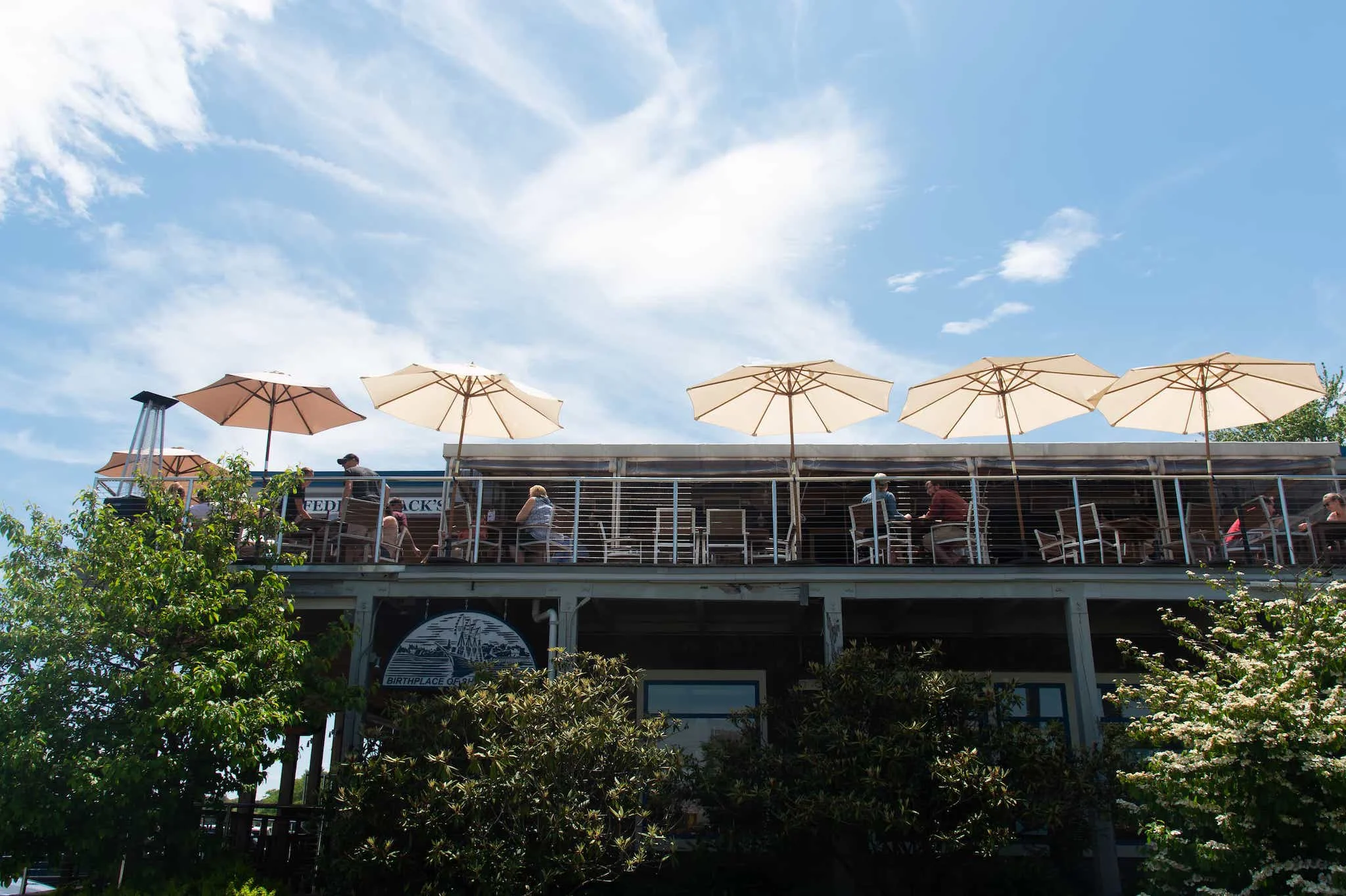 An outdoor dining area with large umbrellas providing shade, filled with patrons enjoying meals under a bright sky.