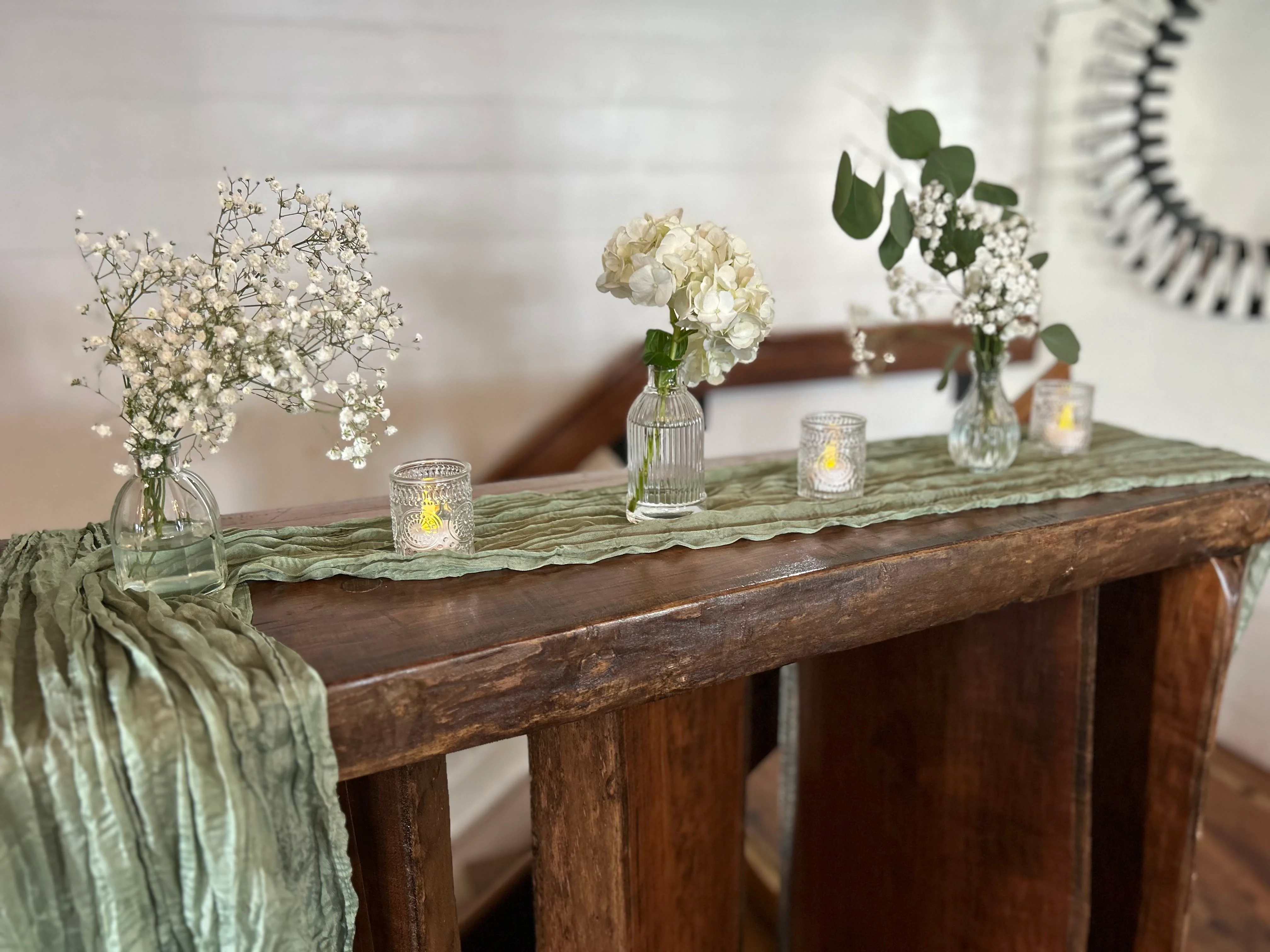 An elegant display of flowers and candles on a wooden surface, featuring baby’s breath and hydrangea in glass vases.