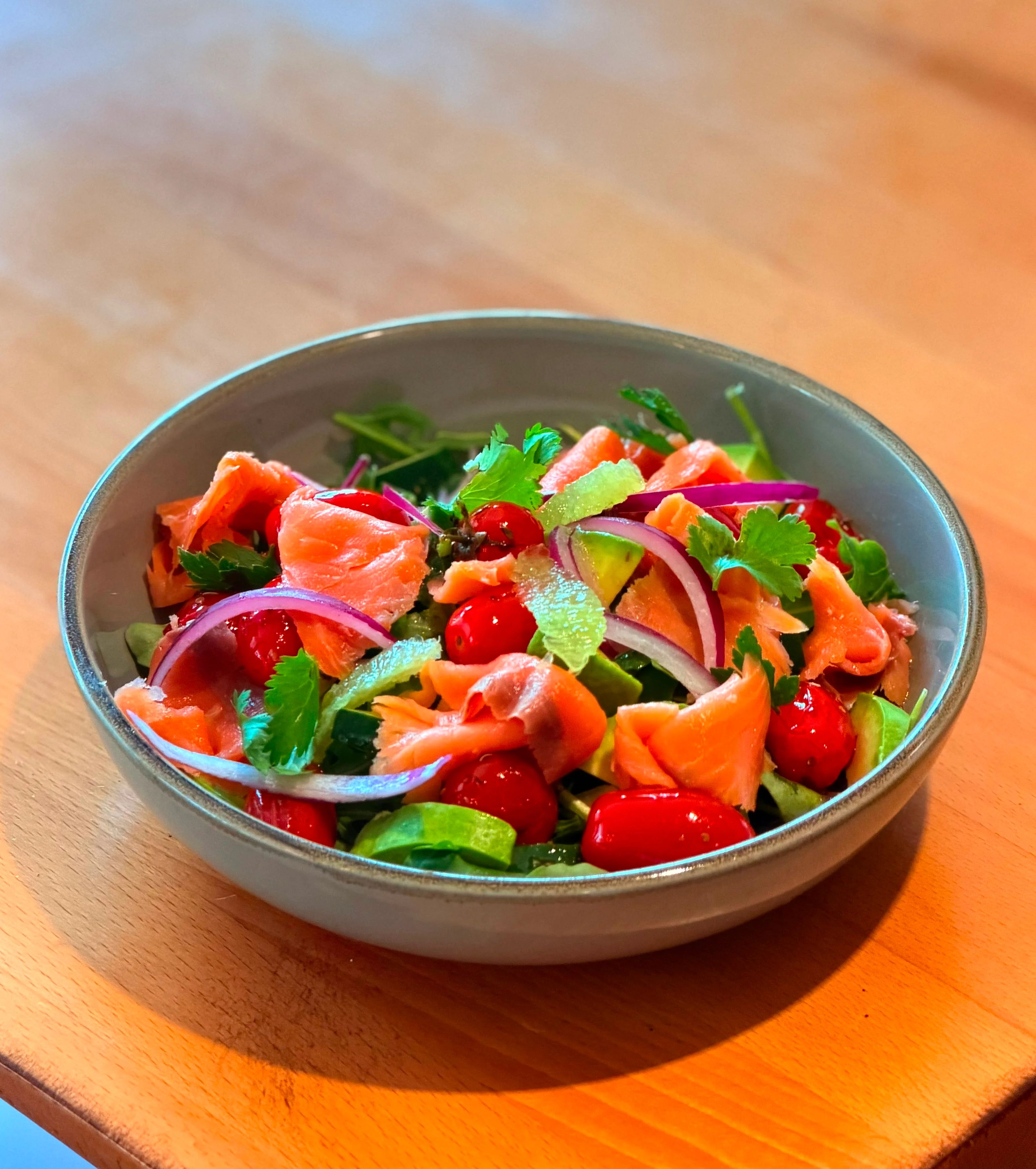 A fresh salad with mixed greens topped with slices of smoked salmon, cherry tomatoes, and red onion, garnished with cilantro.
