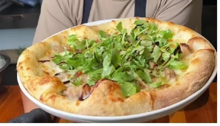 A freshly baked pizza topped with a generous amount of leafy greens, held by a chef. The pizza has a thick, fluffy crust and is presented on a white plate.
