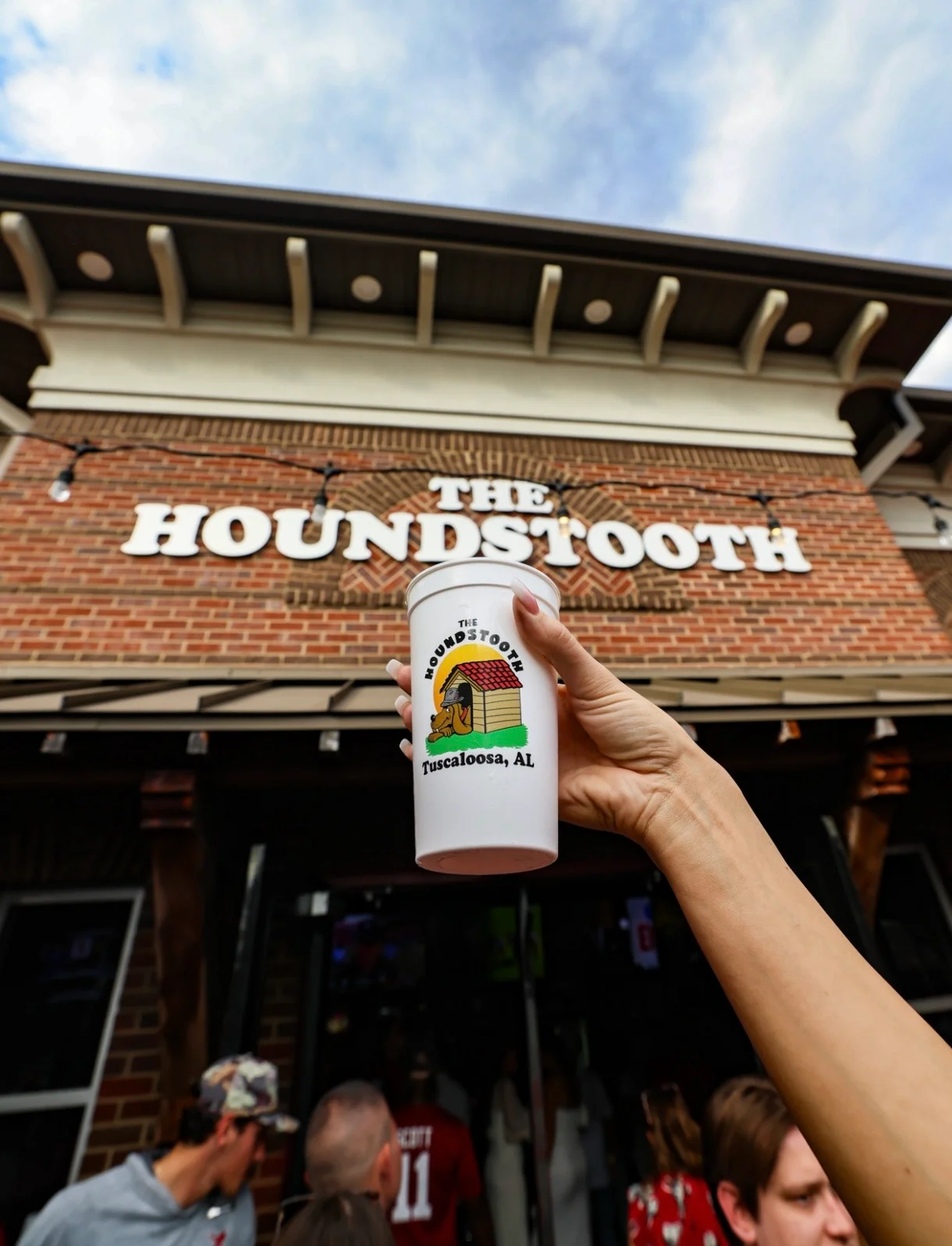 A hand holding a cup in front of The Houndstooth restaurant in Tuscaloosa, AL.