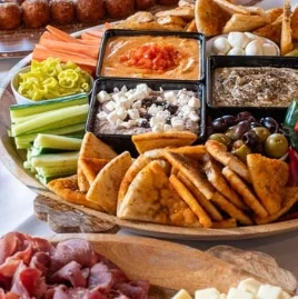 A wooden platter filled with an assortment of appetizers, including pita chips, dips, fresh vegetables, and olives. The arrangement is vibrant and inviting.