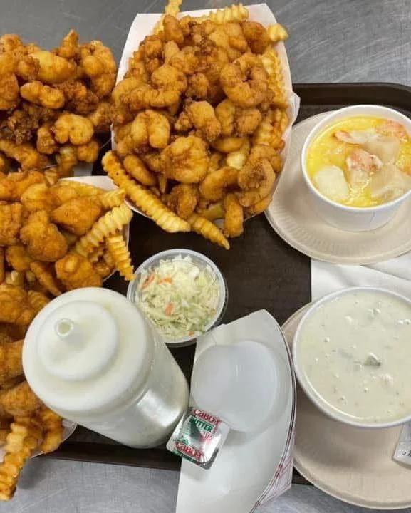 A tray filled with fried shrimp, crinkle fries, a cup of seafood chowder, and coleslaw, accompanied by a condiment container.