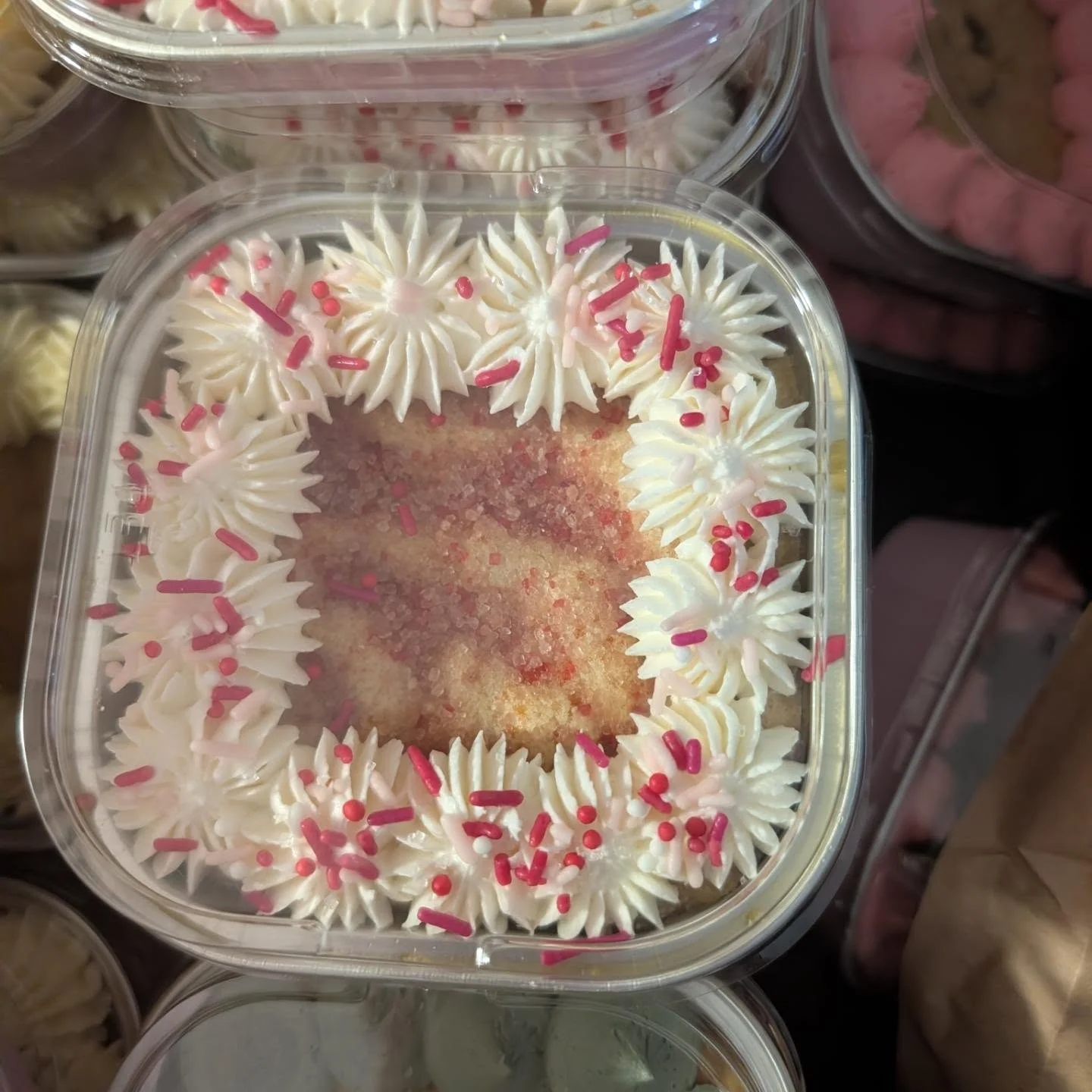 A decorated dessert in a clear container, featuring white frosting with pink and red sprinkles around the edges.