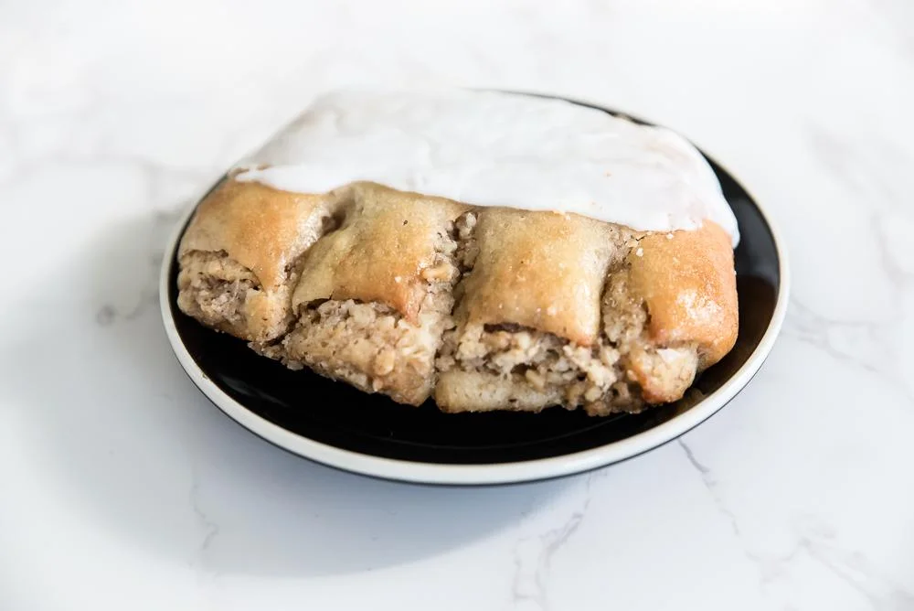 A flaky pastry topped with white icing, showing a textured interior, sits on a black plate with a white rim against a light marble background.