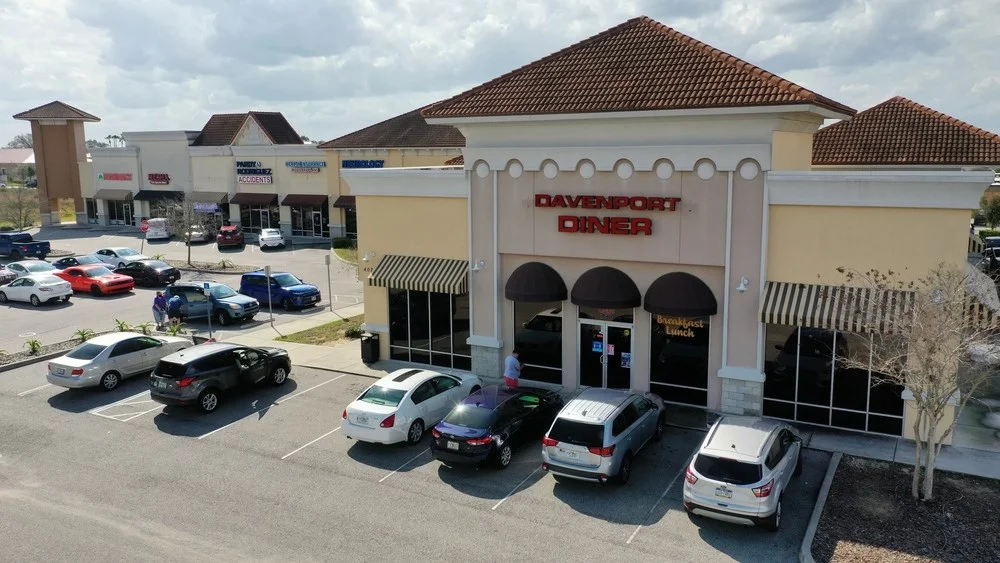 Aerial view of Davenport Diner, a welcoming restaurant with a beige exterior and red signage, located in a strip mall with parked cars in the lot and a partly cloudy sky above.