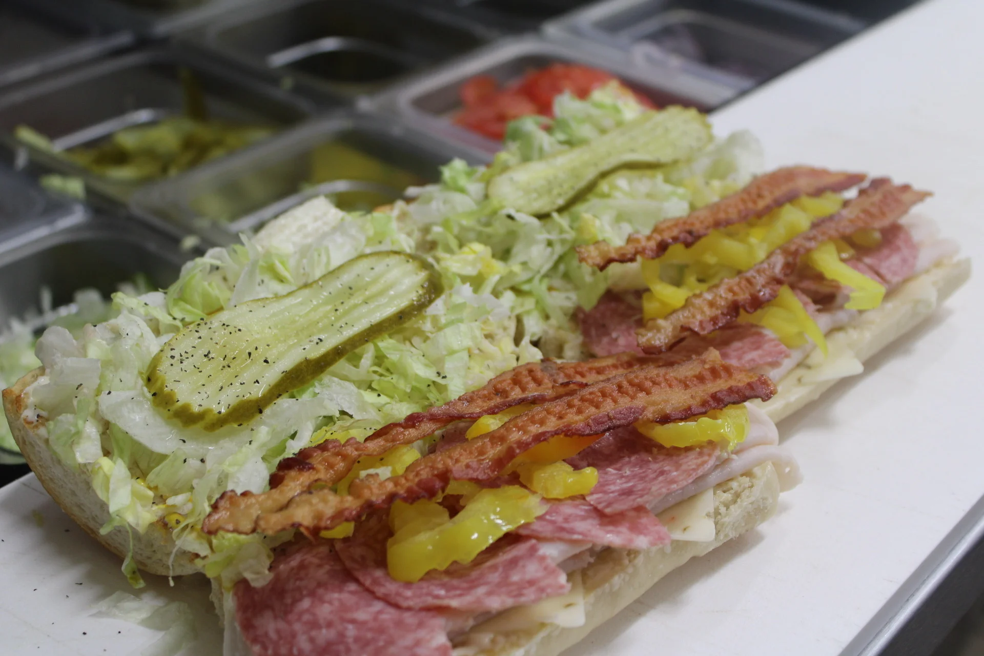 A sub sandwich filled with layers of lettuce, pickles, bacon, and sliced meats, topped with yellow peppers.