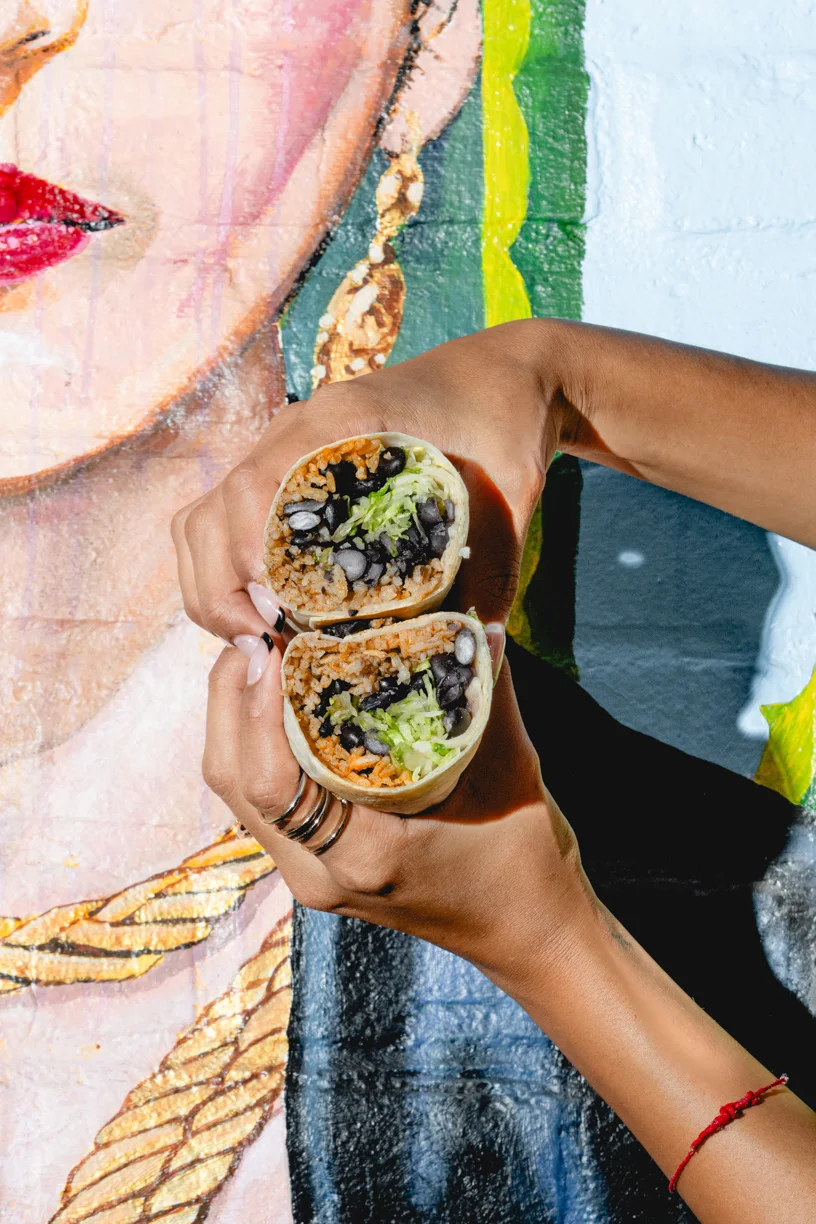 Hands holding a burrito cut in half, filled with rice, black beans, and lettuce in front of a Frida Kahlo mural at Friducha in Brooklyn, NY.
