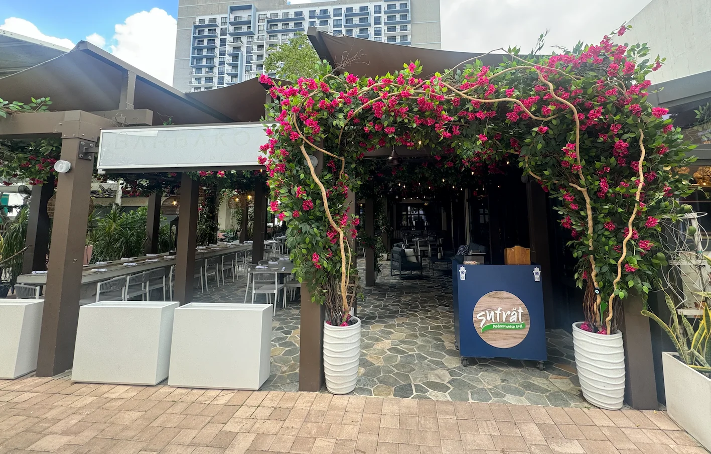 Outdoor restaurant entrance with vibrant pink flowers arching over the doorway. The interior features stone flooring and modern seating. Urban backdrop.