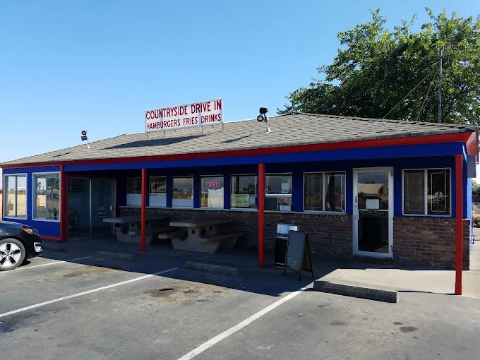Exterior of Countryside Drive-In, a classic diner with a red and blue facade and a vintage-style sign that reads 'Hamburgers, Fries, Drinks.' The building has large windows, outdoor seating with picnic tables, and an open sign welcoming guests. Surrounded by a parking area, the diner exudes a casual, laid-back atmosphere, perfect for enjoying a meal.