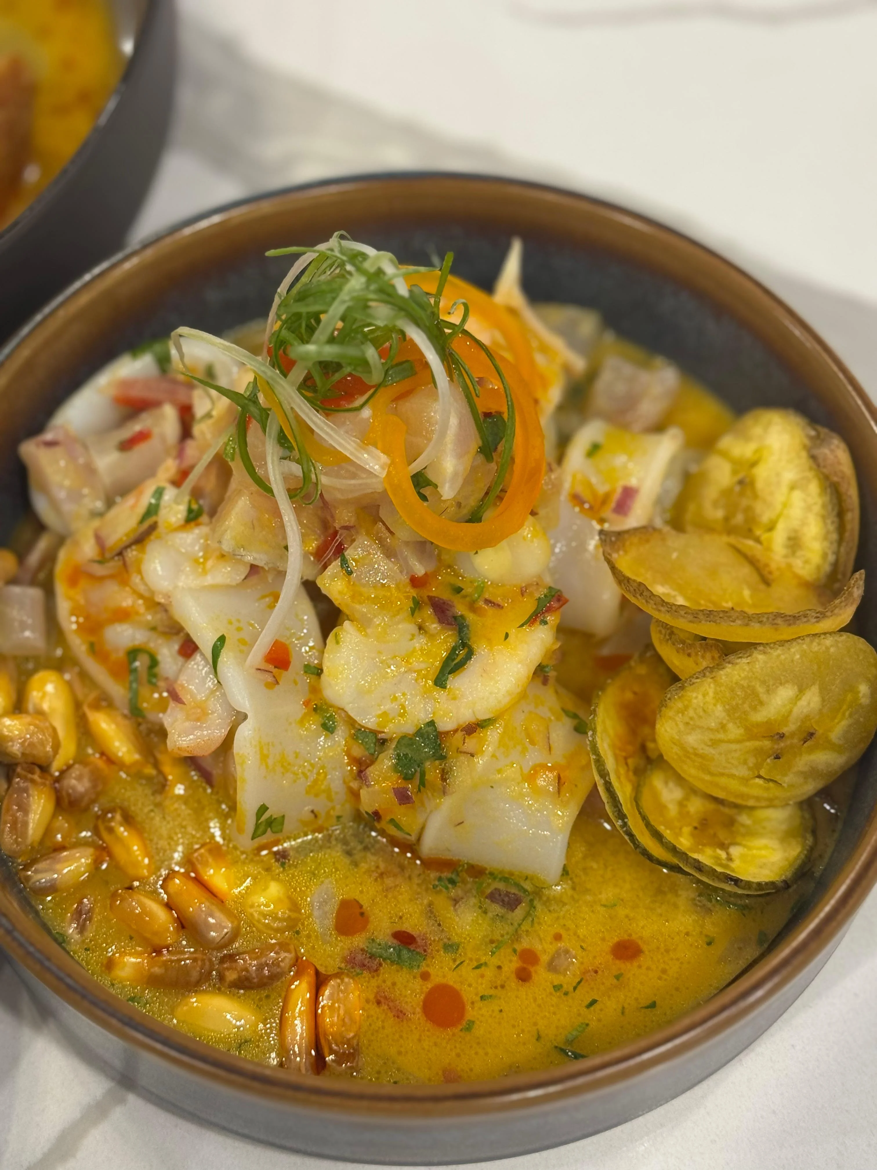 A bowl of seafood dish featuring slices of fish and shrimp in a rich, spicy broth, garnished with herbs and served with crispy banana chips.