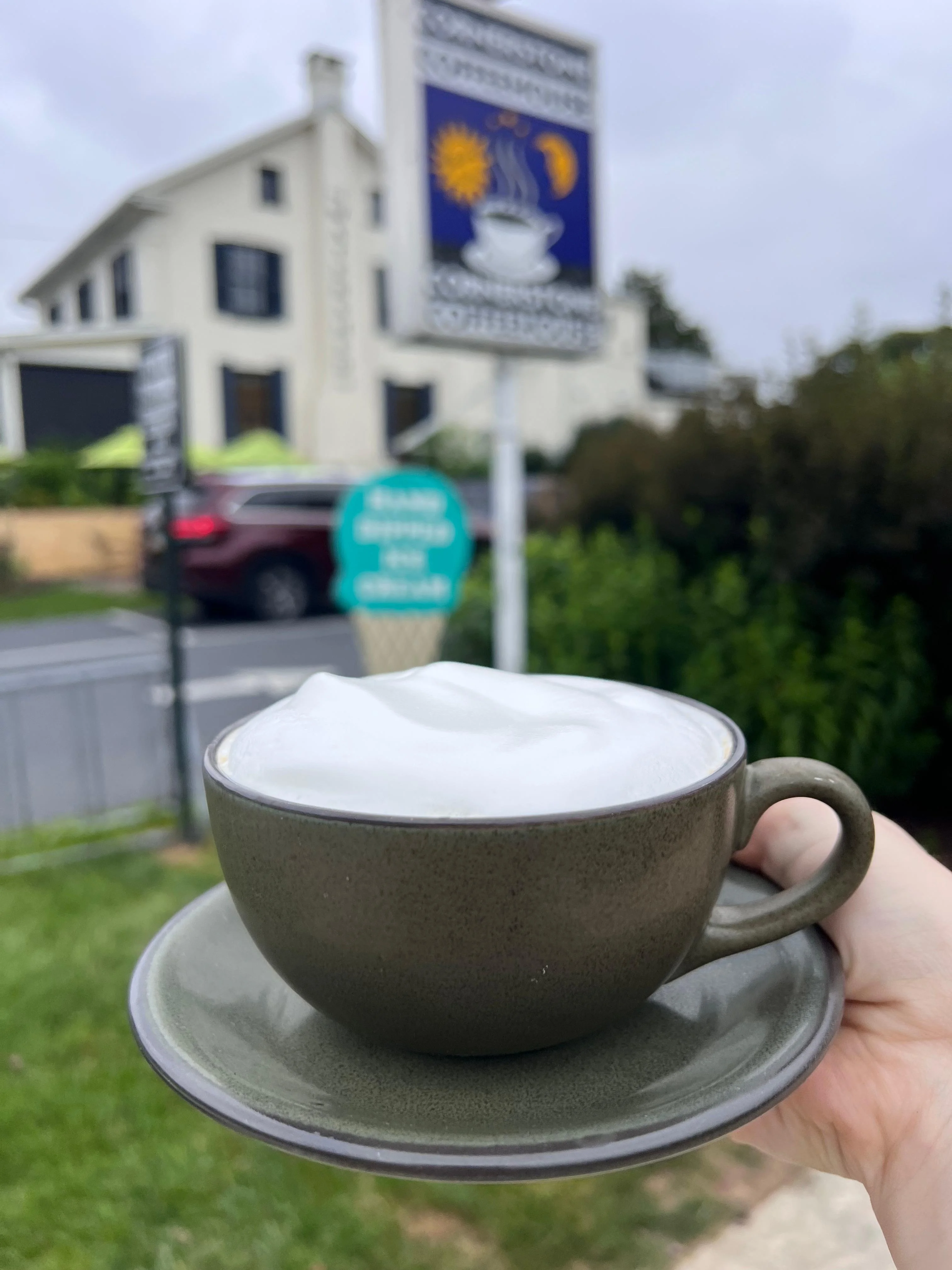 A cup of cappuccino topped with frothy milk, held in front of a coffee shop sign and a green landscape.