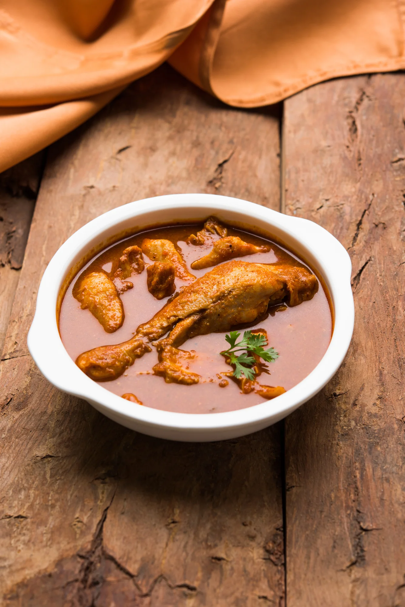 A bowl of chicken curry with tender pieces of chicken in a rich brown sauce, garnished with fresh cilantro. The bowl is placed on a wooden surface with a soft fabric nearby.