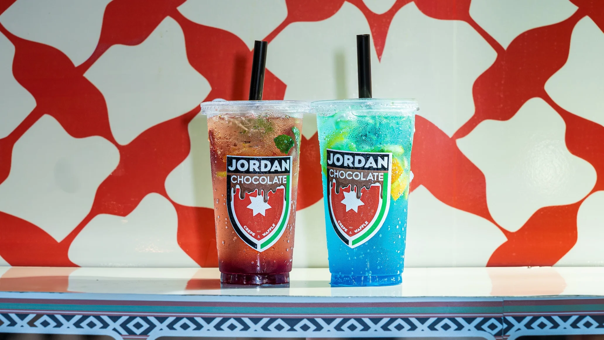 Two colorful drinks in clear cups placed on a table. One is red with lemon and mint, and the other is blue with similar garnishes, both branded with a logo.