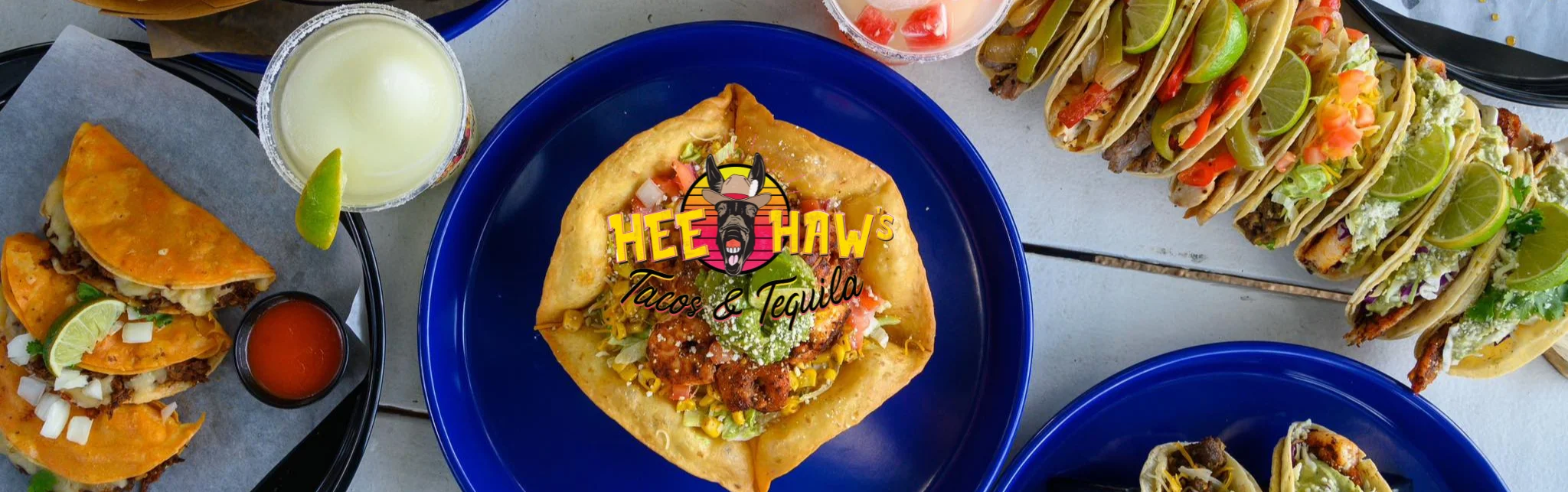 A vibrant spread of tacos and a large taco salad with toppings, accompanied by drinks and garnishes.