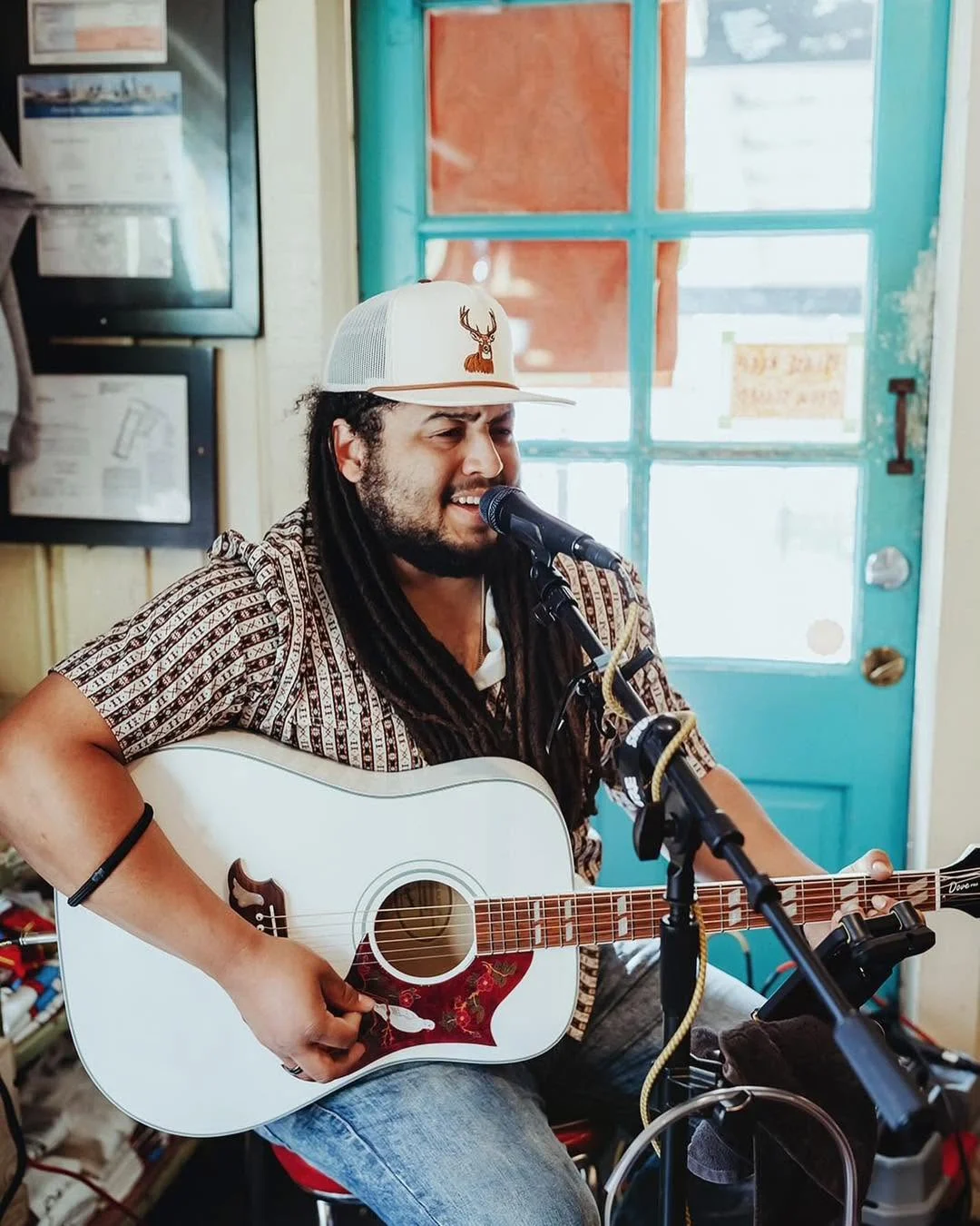 A performer playing an acoustic guitar inside a cozy venue, singing into a microphone. The background features a blue door and various framed documents.
