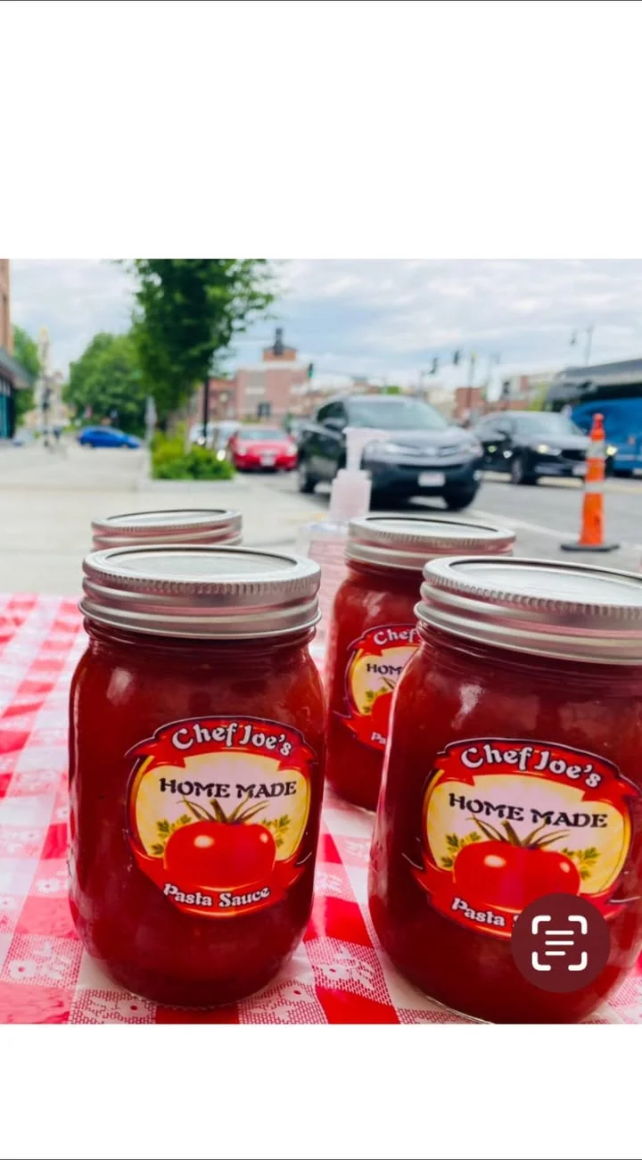 Jars of homemade pasta sauce are neatly arranged on a table, featuring colorful labels, in an outdoor setting.