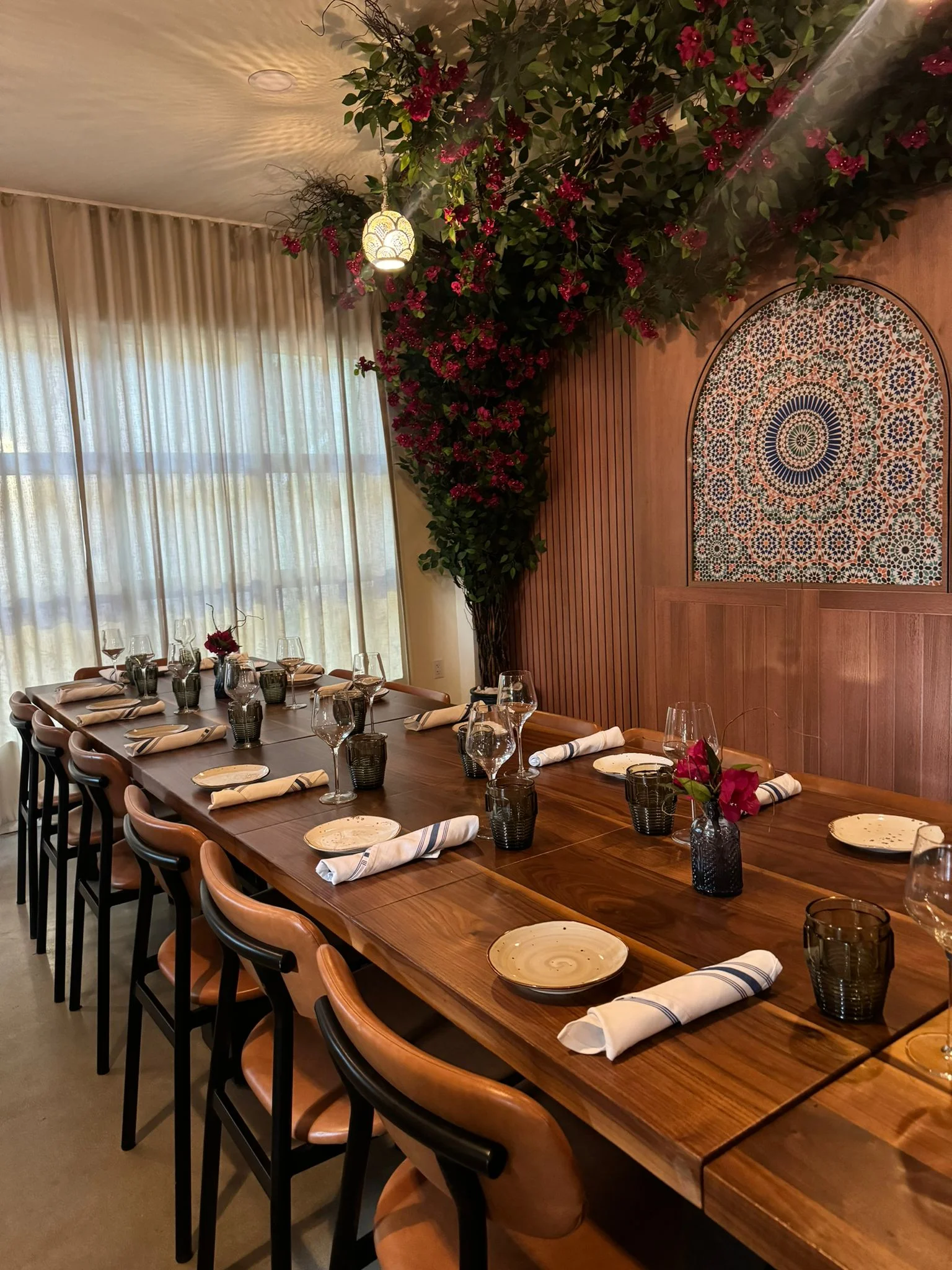 Elegant dining room featuring two long wooden tables set with plates, glasses, and napkins. Ornate tile paneling and hanging lights create a cozy atmosphere.
