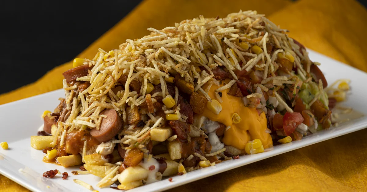 A large serving of loaded nachos featuring fries topped with a variety of ingredients including hot dogs, cheese, corn, and crispy fried onions. The dish is presented on a white platter against a vibrant yellow background.