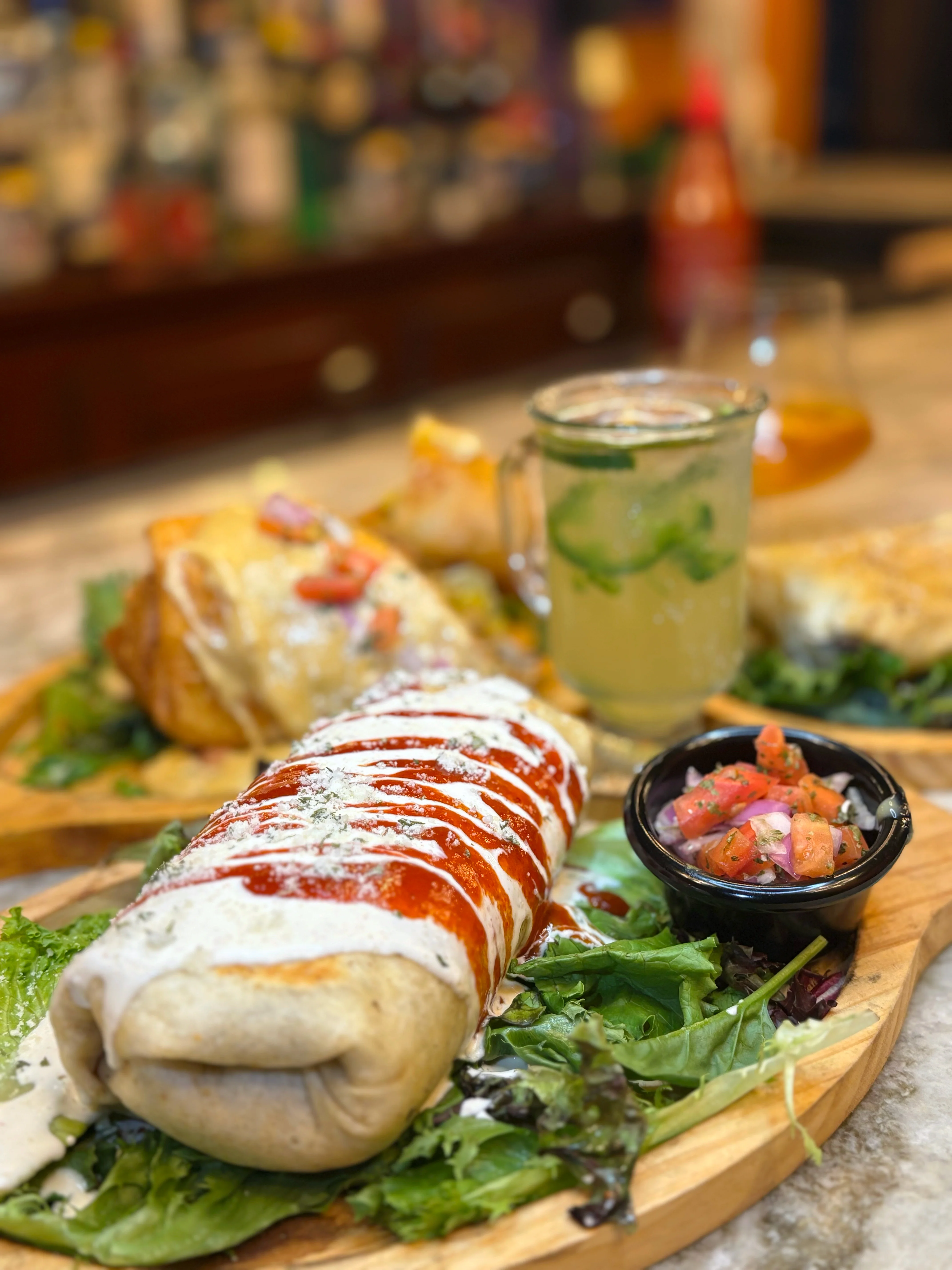 A large burrito drizzled with sauce and served alongside a salad and a refreshing drink. The plate showcases a variety of food items in a vibrant and inviting setting.