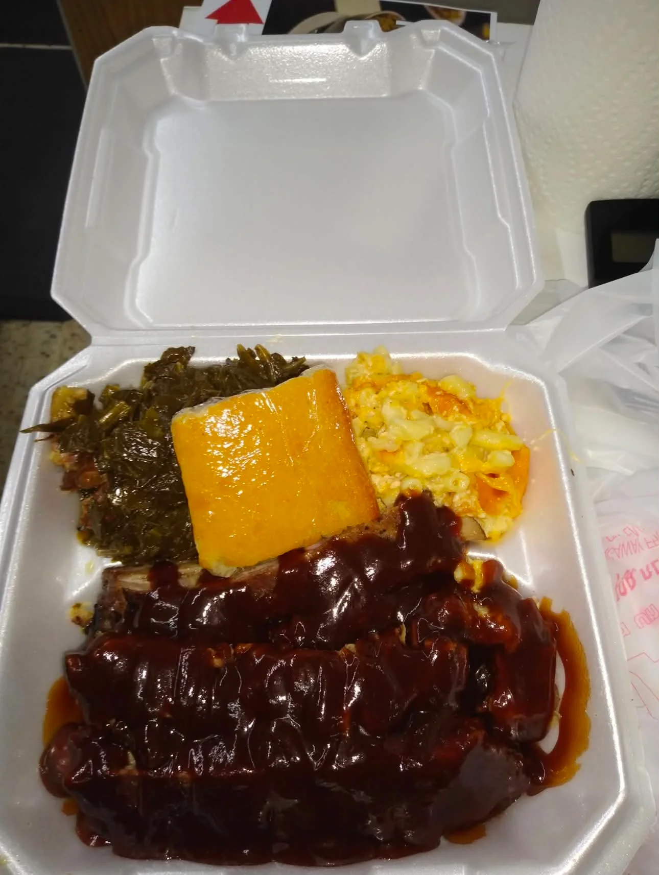 A takeout container filled with barbecue ribs, mac and cheese, greens, and cornbread.