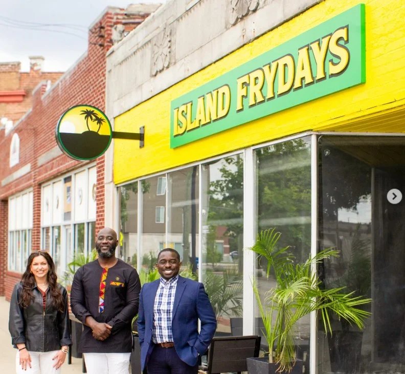 Three individuals standing outside a restaurant named 'Island Frydays'. The restaurant features bright yellow walls and palm tree decor.
