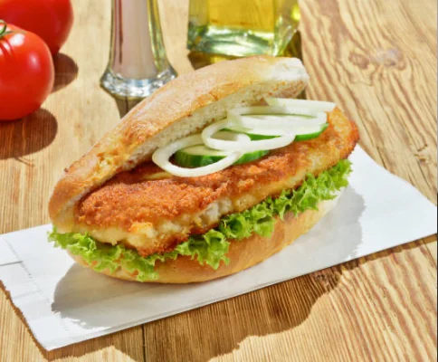 A crispy fish sandwich served on a toasted bun, topped with lettuce, fresh onions, and garnished with tomatoes in the background.
